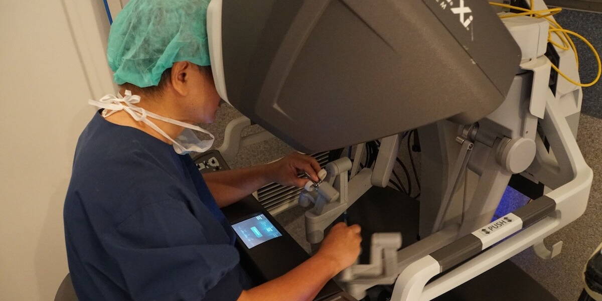 Een chirurg, met een blauw operatieschort en een groen mutsje, zit bij de console van een Da Vinci Xi robotchirurgiesysteem.