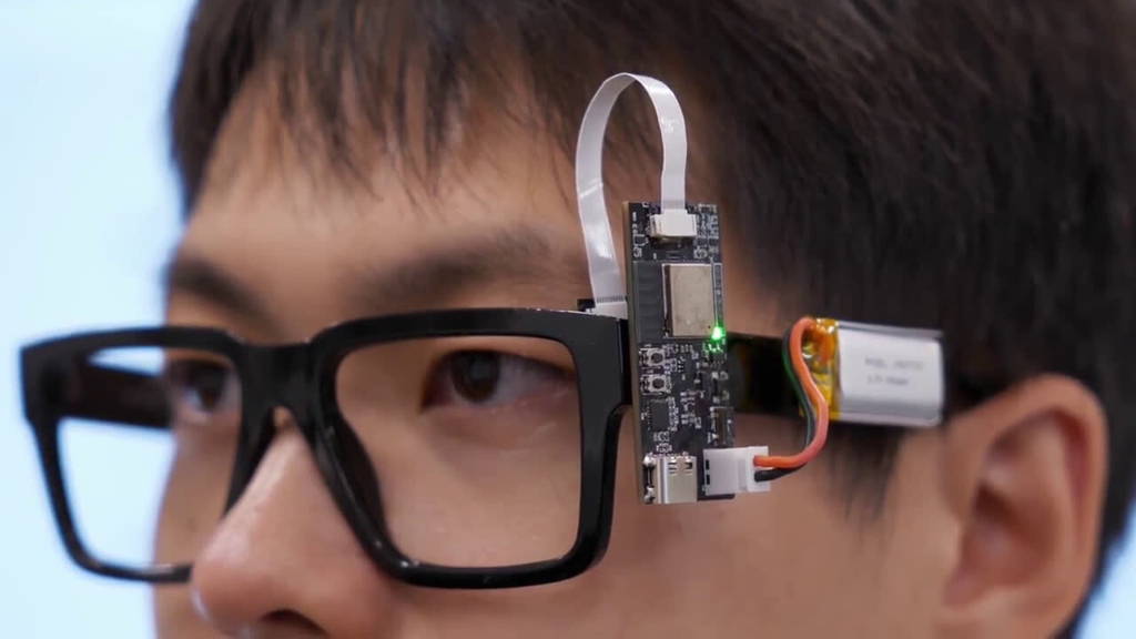 Een close-up toont een persoon die een prototype van een draagbare computer of sensor draagt, bevestigd aan een zwart montuur van een bril.