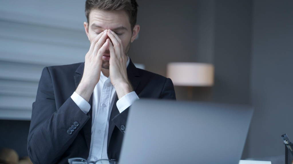 Overworked male entrepreneur sits at home office, suffering from heavy headache and fatigue