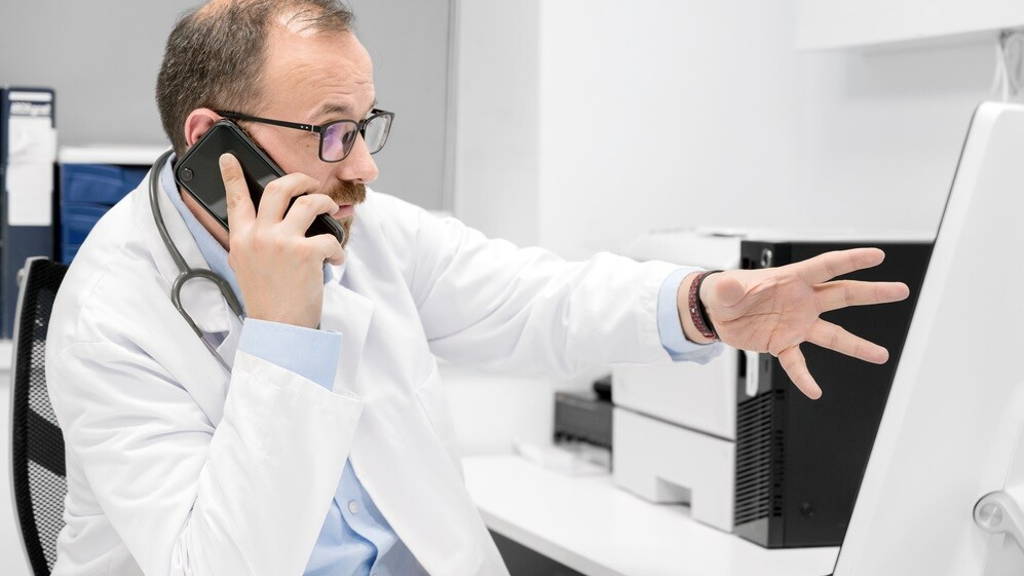 Male doctor pointing with finger on desktop computer while talking on the phone, discussing treatment with colleague.