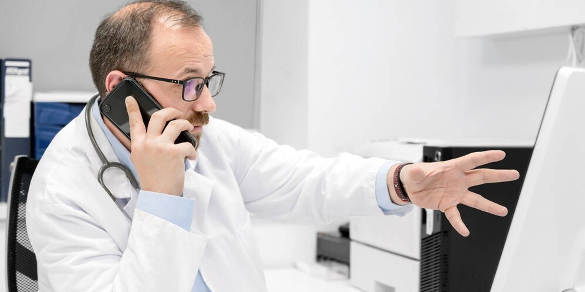 Male doctor pointing with finger on desktop computer while talking on the phone, discussing treatment with colleague.