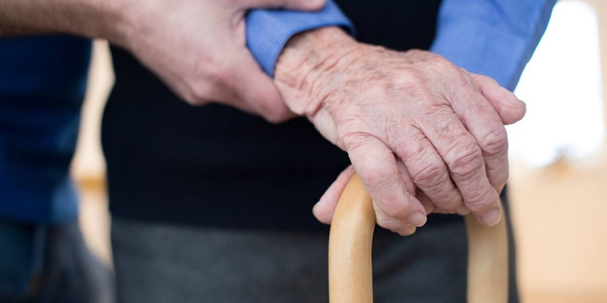 Senior Man's Hands On Walking Stick With Care Worker In Background