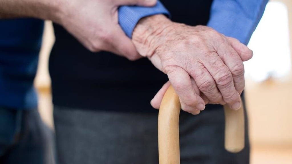 Senior Man's Hands On Walking Stick With Care Worker In Backgrou