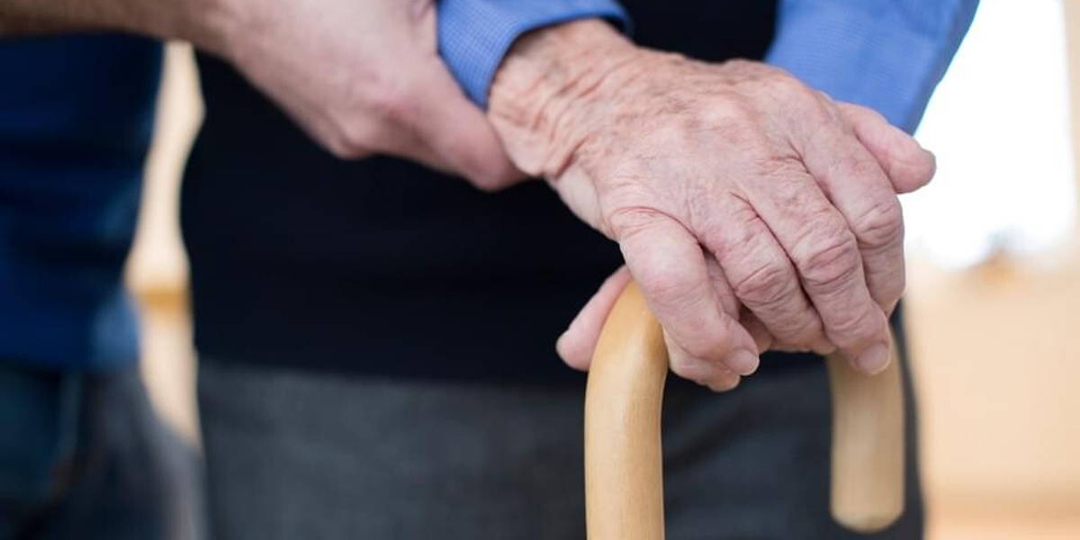 Senior Man's Hands On Walking Stick With Care Worker In Backgrou